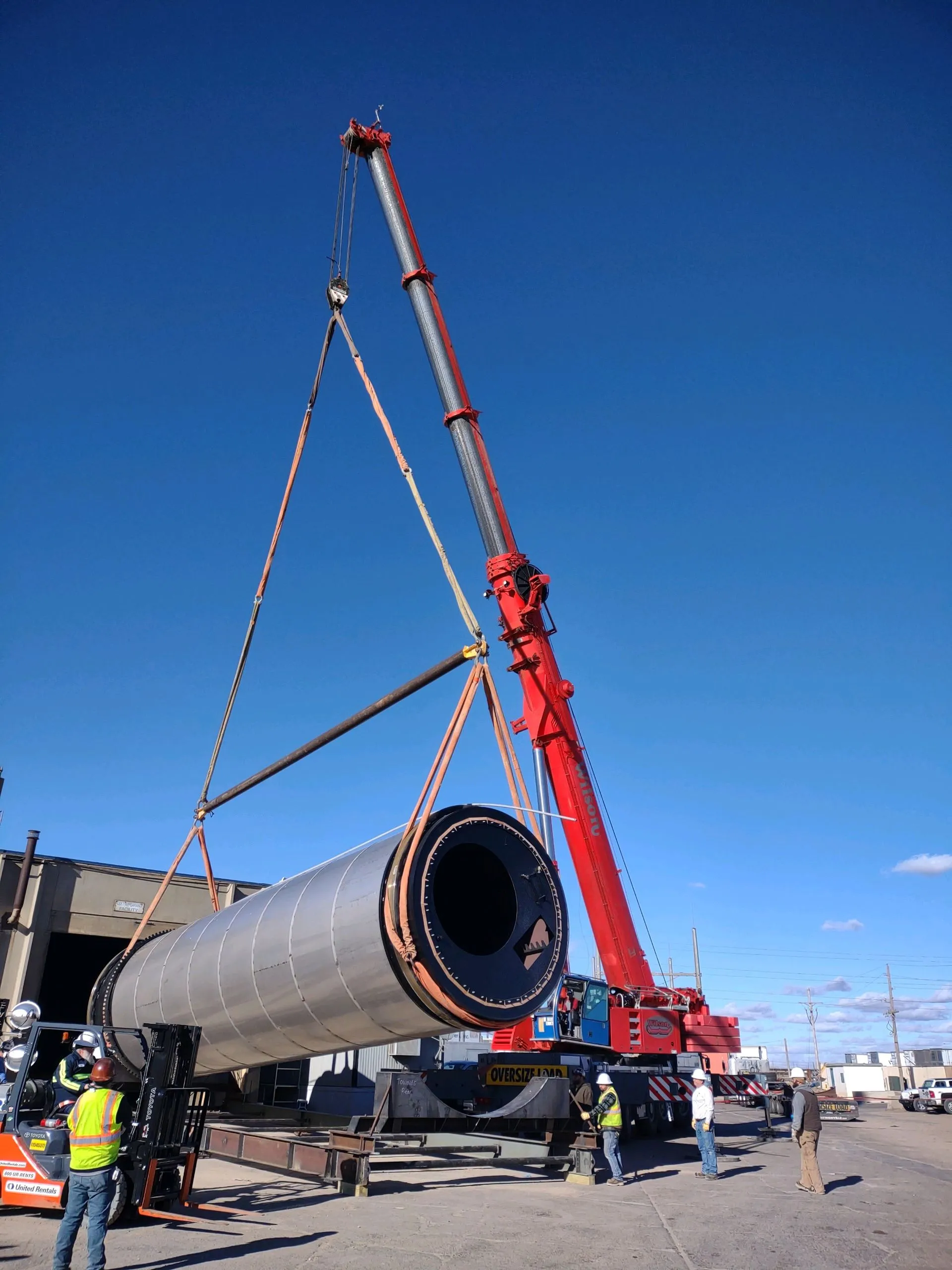 Red all-terrain crane raising a large steel cylinder on slings and a spreader bar under a clear blue sky