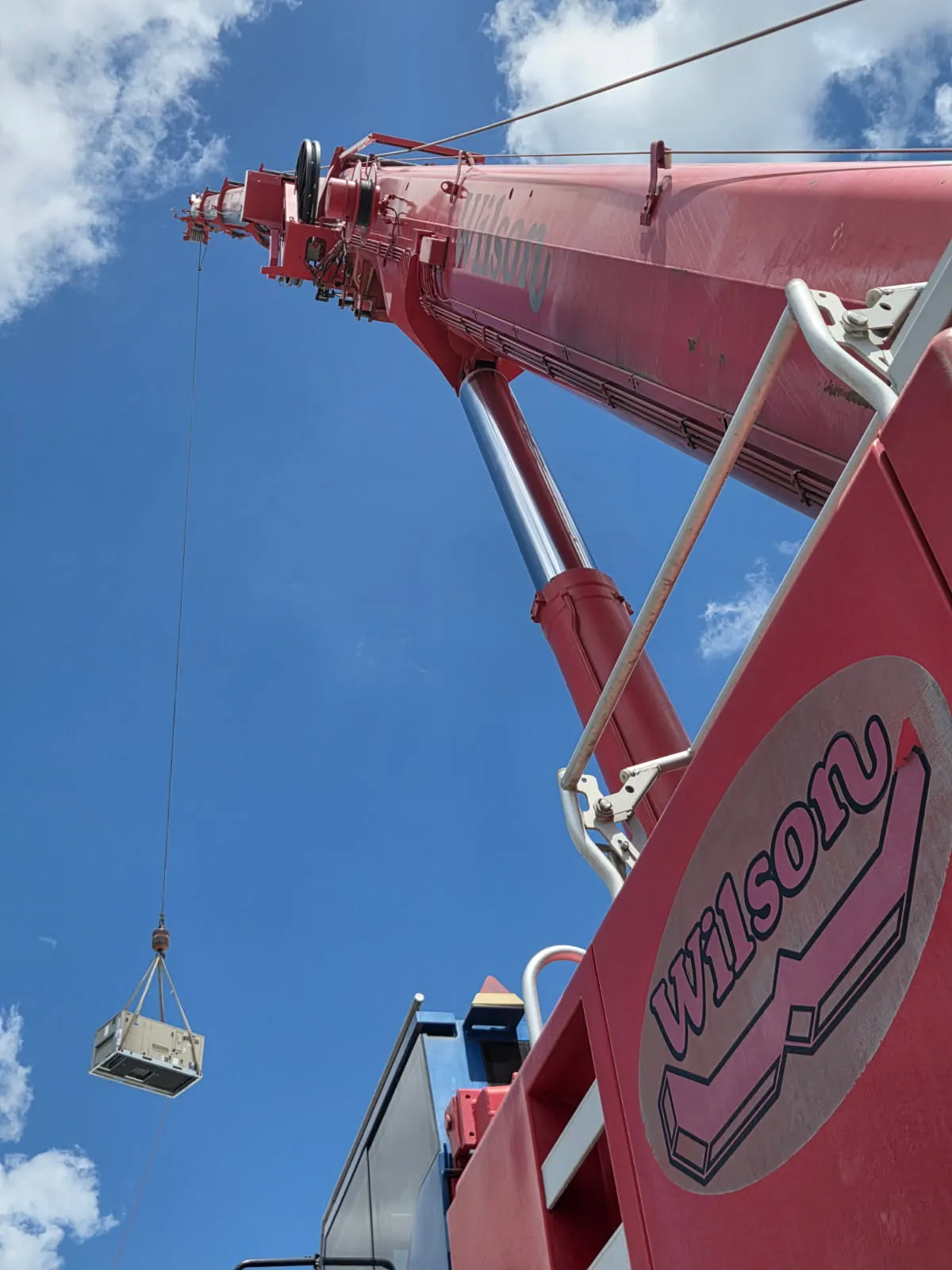 Crane boom lifting a rooftop HVAC unit into the air against a blue sky