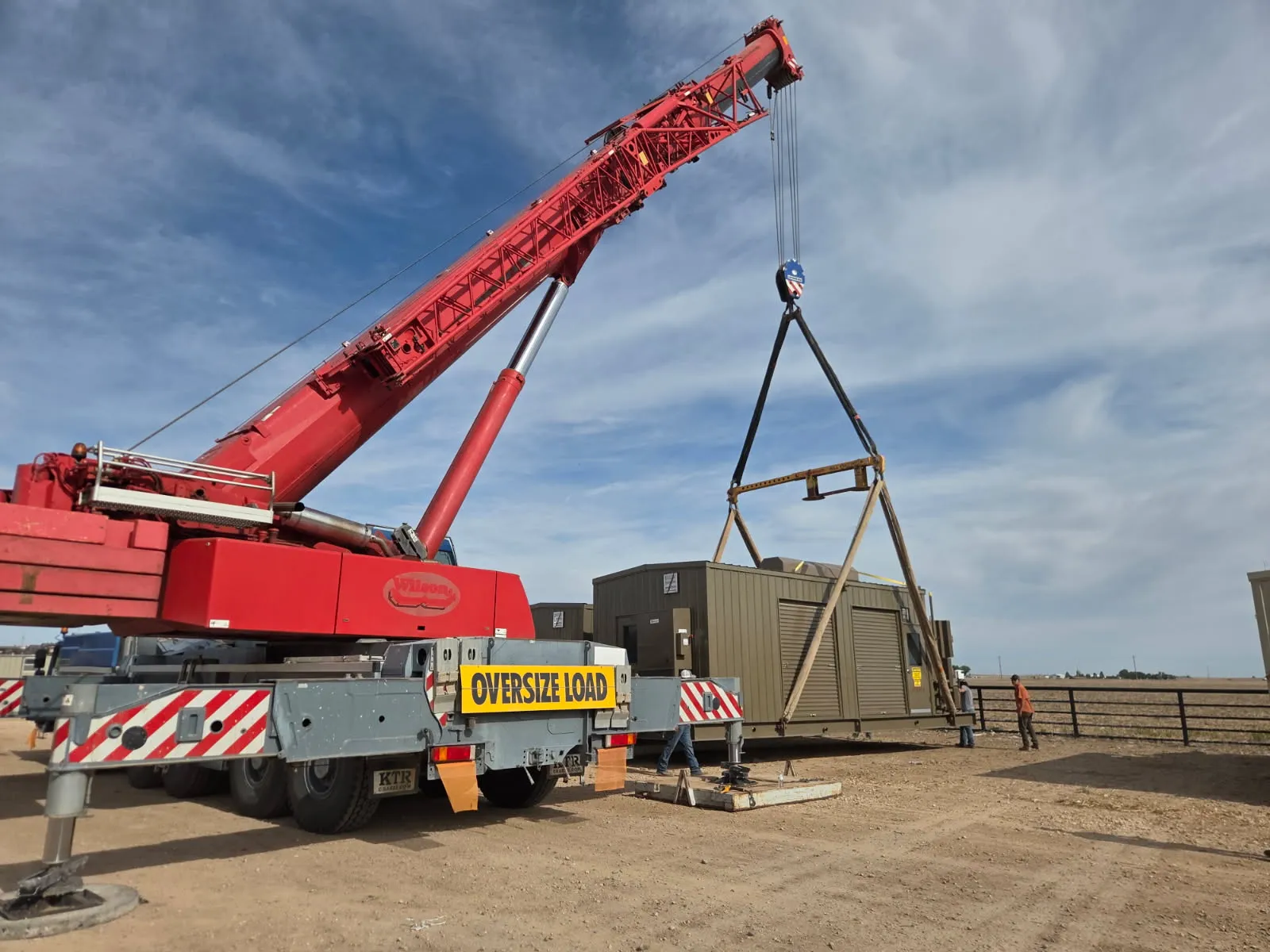 Jim Wilson Crane Service crane setting a large equipment building onto its foundation at a rural site, with the crew standing by
